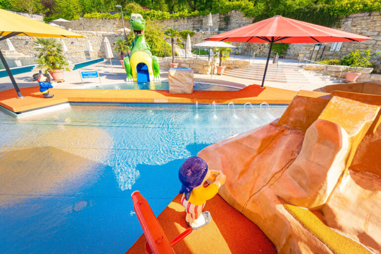 Blick über das bunte Kinder-Planschbecken mit Rutsche, Piratenfigur und großem Drachen im Hintergrund, umgeben von Sonnenschirmen und Steinmauern.