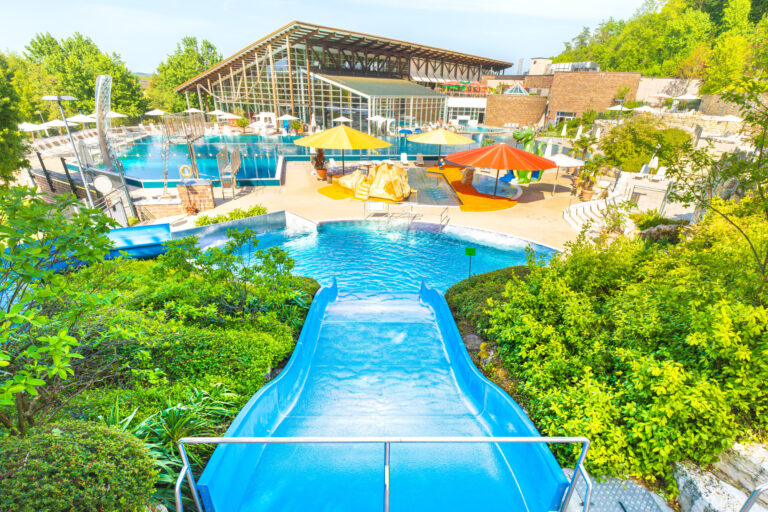 Zwei große blaue Rutschen führen in ein separates Becken, umgeben von terrassenartig angelegter Gartenlandschaft mit Natursteinmauern.