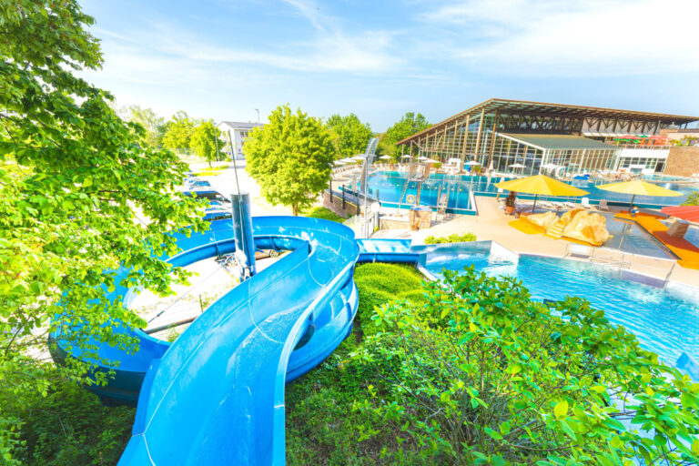 Luftaufnahme auf ein Freibad mit mehreren Becken, Rutschen, Sonnenschirmen und vielen Liegeflächen.