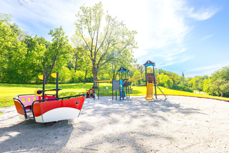 Spielplatz, der im Thema Pirat gehalten ist mit Schaukeln und Rutschen auf Kiesboden, eingebettet in eine grüne Parklandschaft.