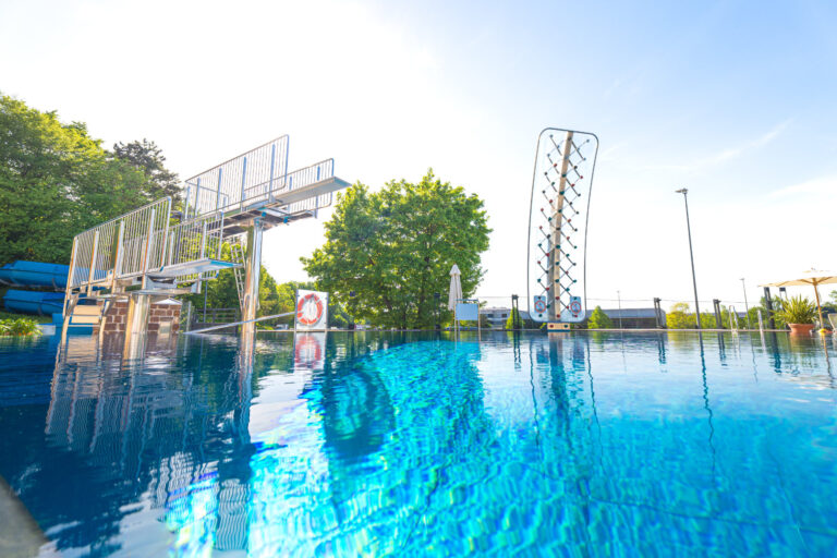 Sprungturm und Kletterwand am Beckenrand des Freibads bei strahlendem Sonnenschein, umgeben von Bäumen und Liegen.