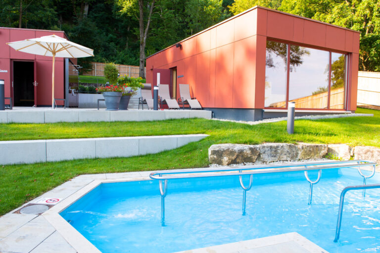 Kneipbecken vor einem modernen roten Saunahaus mit Sonnenschirmen und Liegestühlen auf grünem Rasen.