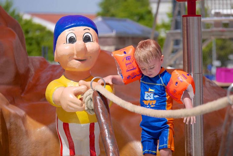 Kleinkind mit Schwimmflügeln im bunten Badeanzug neben einer Piratenfigur im Kinder-Erlebnisbereich des Freibads – beide halten sich an einem Seil fest.