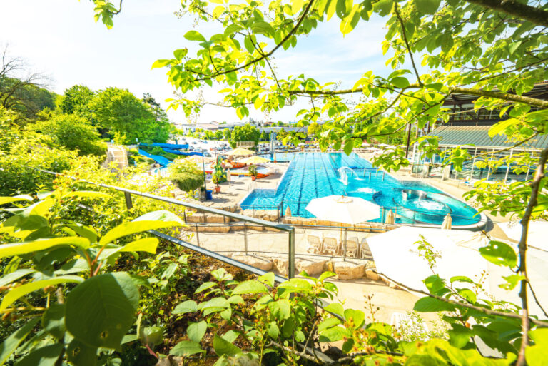 Panoramaaufnahme des Freibads mit Schwimmbecken, Sonnenschirmen und Rutschen, eingerahmt von grüner Natur – Blick von einem höher gelegenen Weg.