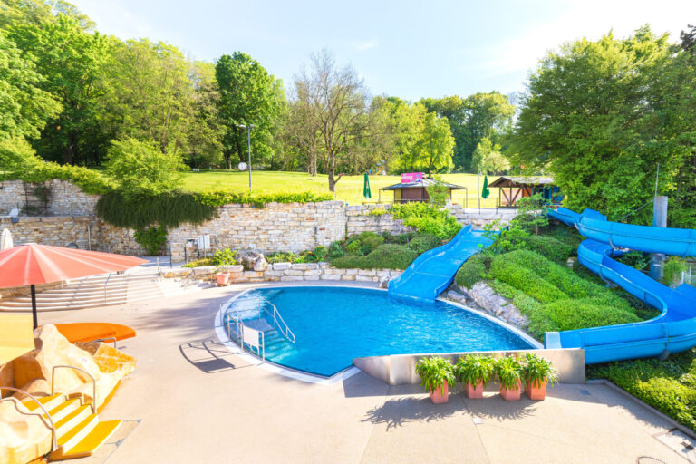 Zwei blaue Rutschen führen in ein separates Becken, umgeben von grüner Hanglandschaft und einer Natursteinmauer.