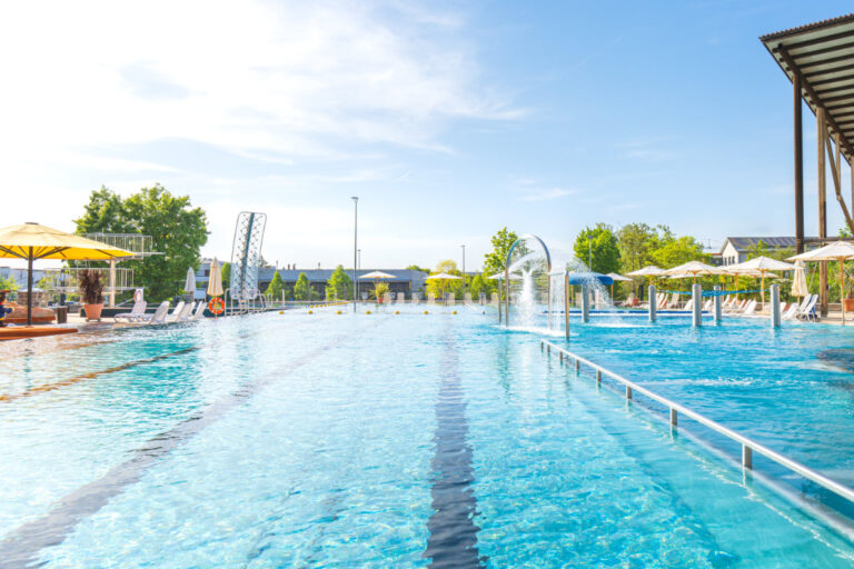 Langes Schwimmerbecken mit Startblöcken, Wasserattraktionen und schattenspendenden Schirmen.