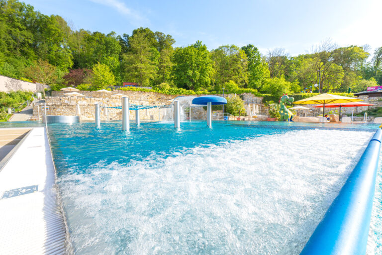 Sprudelbecken mit verschiedenen Wasserdüsen und Fontänen, umgeben von Natursteinmauer, Bäumen und Sonnenschirmen.