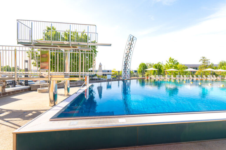 Sprungturm mit Kletterwand am klaren Schwimmbecken unter freiem Himmel – eingerahmt von Liegestühlen und Sonnenschirmen.