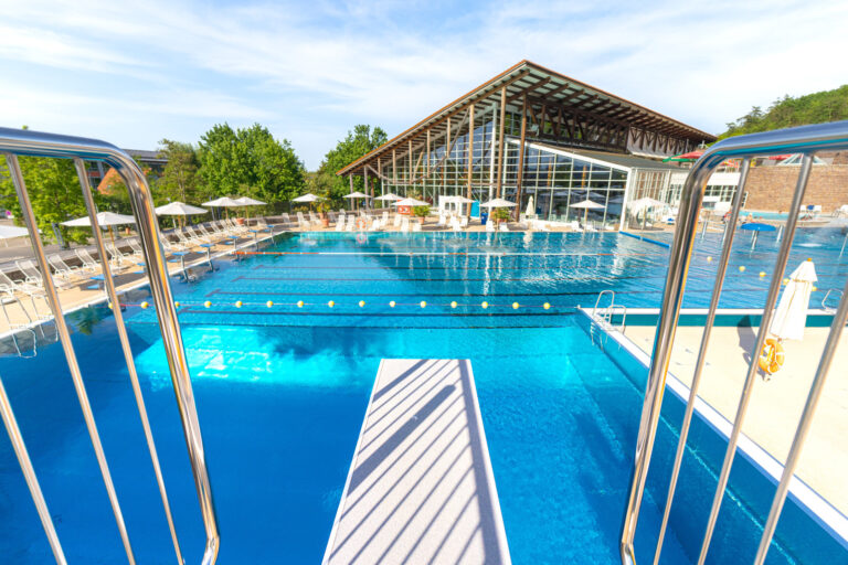 Blick vom Sprungturm auf das große Freibadbecken mit markierten Bahnen, Liegestühlen und dem verglasten Hallenbadgebäude.